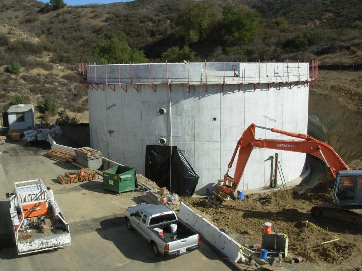 Construction site with a large concrete cylindrical structure, excavator, vehicles, and building materials.