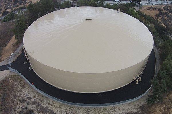 A large, beige circular storage tank sits in a fenced area surrounded by trees and hills.