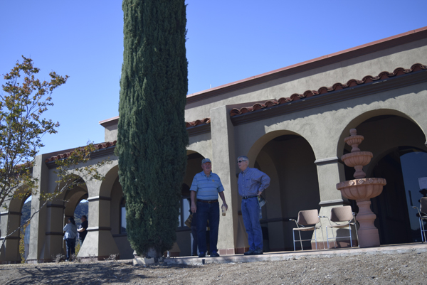 Two people standing outside a building with arches, near a tall tree and a fountain.