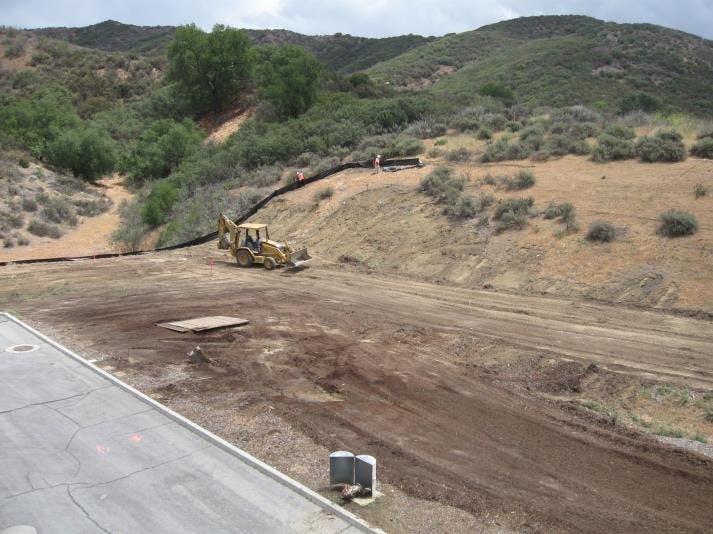 A construction site with a bulldozer working on a hillside, surrounded by greenery and dirt. Some workers are visible.