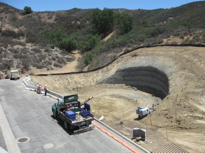 Construction site with trucks, workers, and excavated hole in a hilly area.