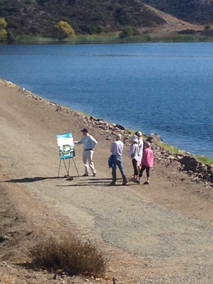 Four people stand by a lake, looking at a poster on an easel on a gravel path.