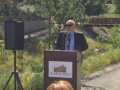 A person speaks at a podium labeled "City of Agoura Hills" outside, with greenery and a bridge in the background.