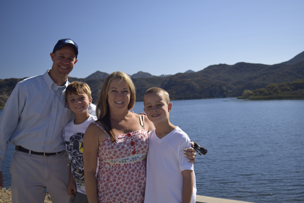 A family of four smiling in front of a lake with mountains in the background.