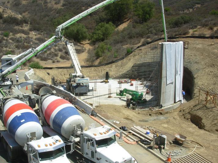 Construction site with cement mixers, a crane, and workers. Concrete being poured into a wall structure surrounded by hills.