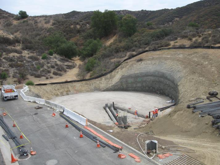 Construction site with a large, circular excavation, surrounded by hills. Workers and equipment are visible.