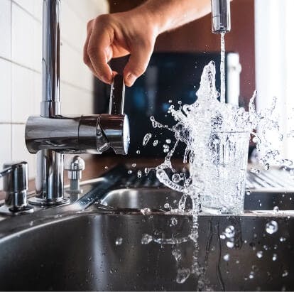 A hand adjusts a faucet while water fills a glass in a sink, causing a splash.