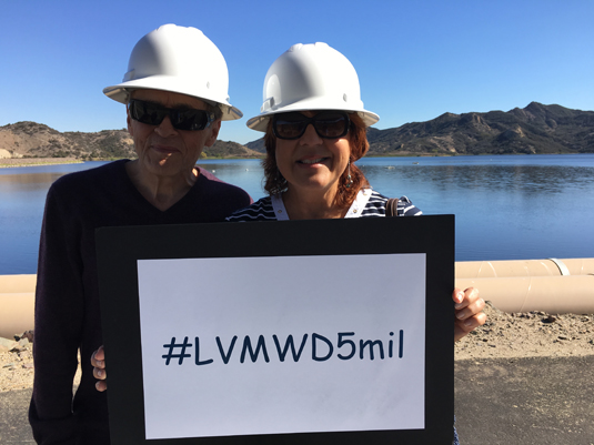 Two people in hard hats hold a sign reading "#LVMWD5mil" by a lake.