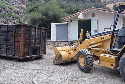 A bulldozer near a rusty dumpster in front of a building with greenery in the background.