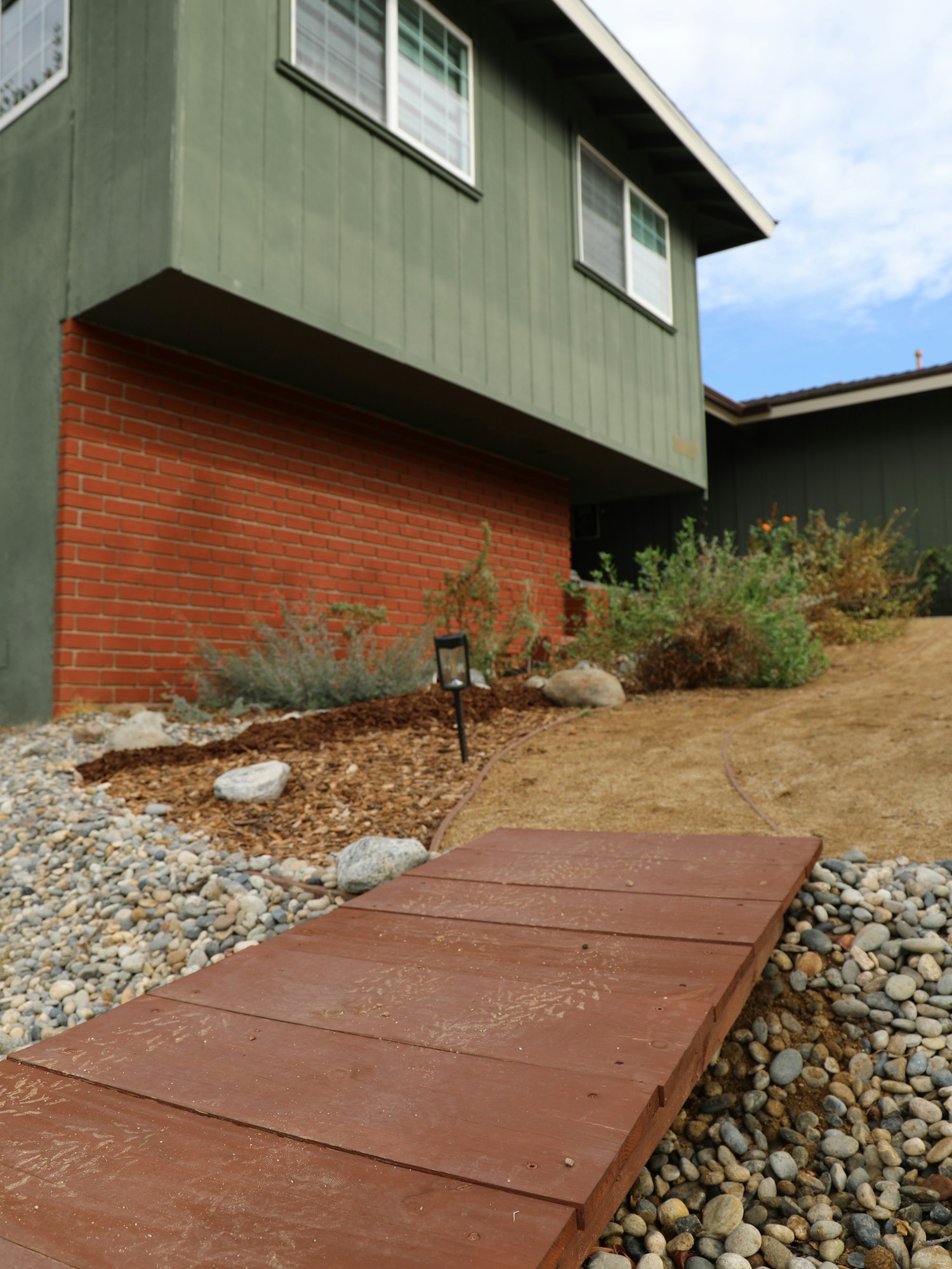 A wooden pathway leads to a house with green siding and a brick base, surrounded by gravel and plants in a landscaped garden.