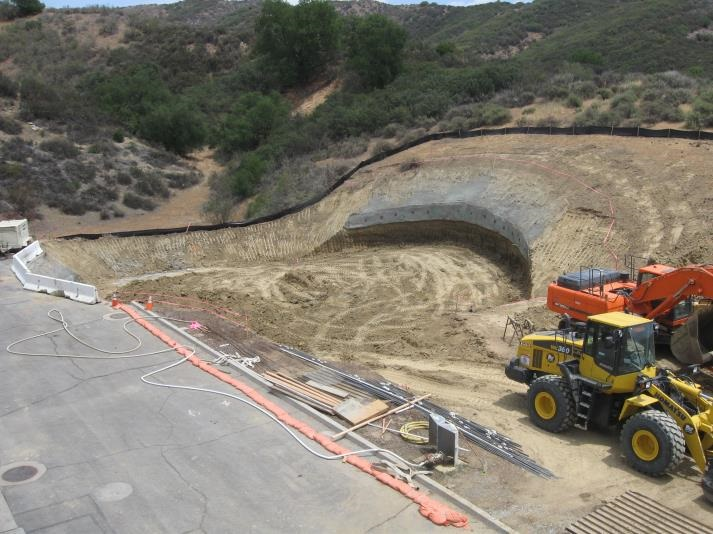Construction site with excavation, heavy machinery, and surrounding hills.