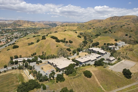 Aerial view of buildings surrounded by hills and sparse trees under a partly cloudy sky.