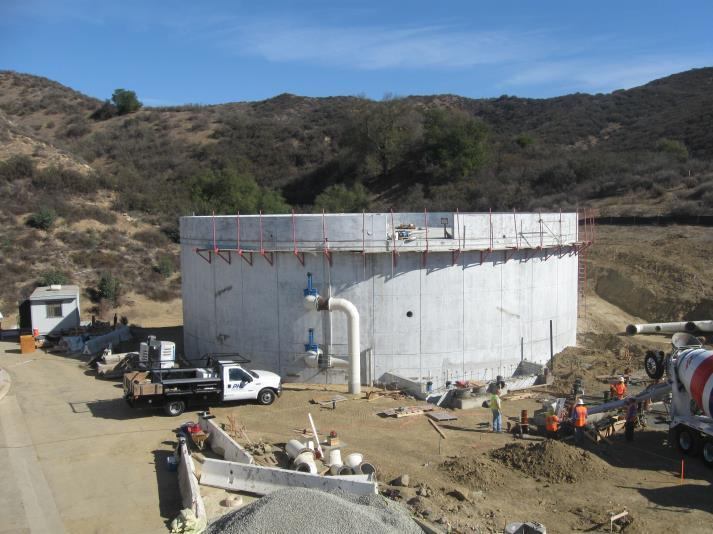 Construction site with a large circular concrete structure, trucks, workers, pipes, and equipment in a hilly area.