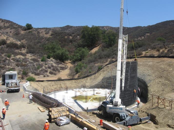 A construction site with workers, a crane lifting materials, and a circular foundation amidst hilly terrain.