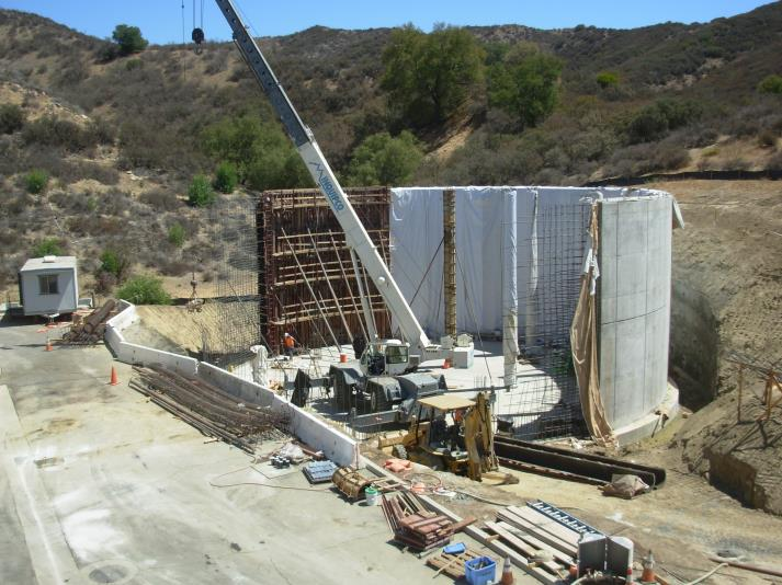 Construction site with a crane building a circular concrete structure in a hilly area. Construction materials are scattered around.