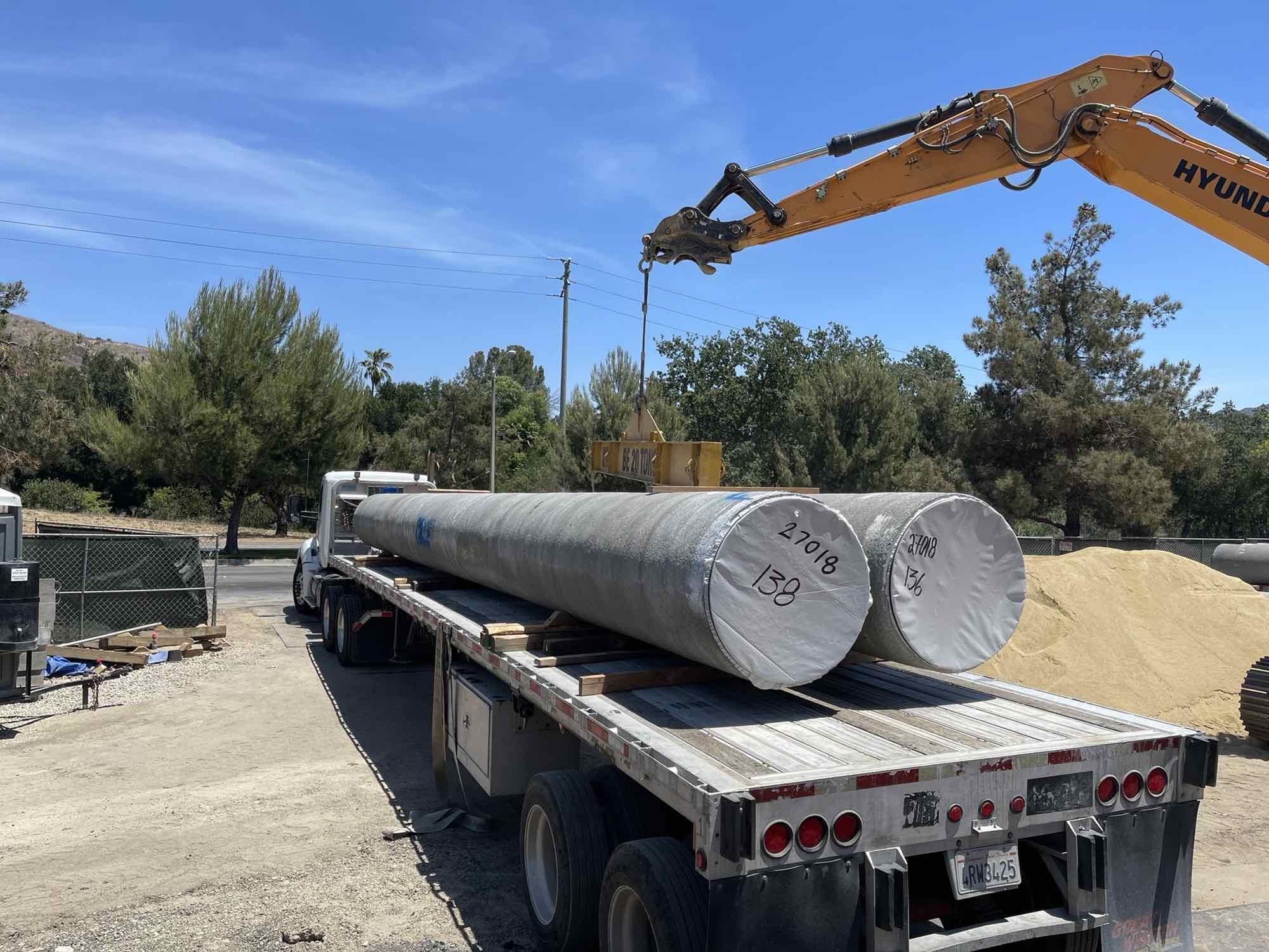 A flatbed truck with large cylindrical items, lifted by a Hyundai excavator, on a construction site with trees in the background.