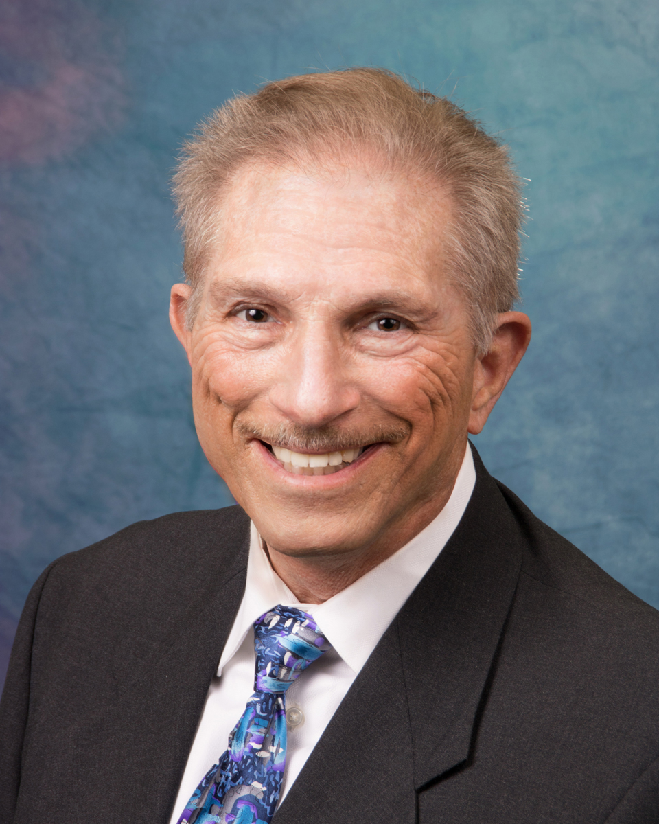 A smiling man in a suit with a colorful tie, set against a blue background.