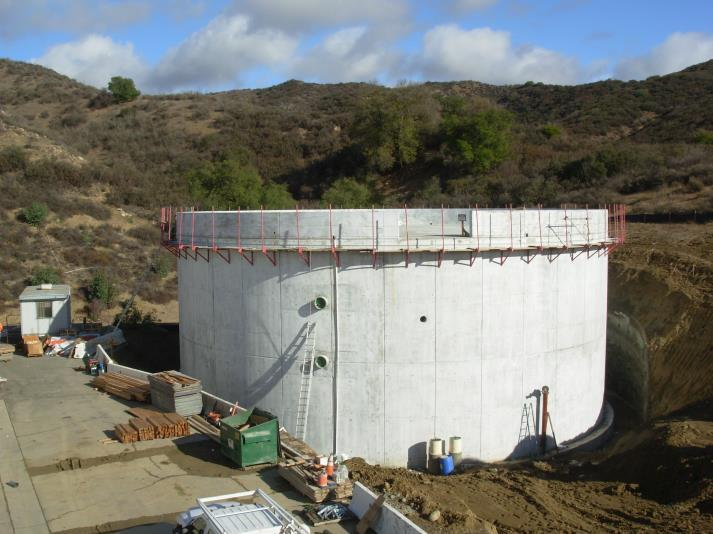 A large concrete structure under construction in a hilly area, surrounded by equipment and construction materials.