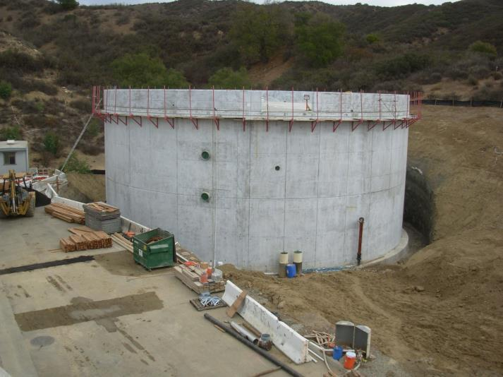 A large, cylindrical concrete structure under construction, surrounded by tools, materials, and equipment in a hilly area.