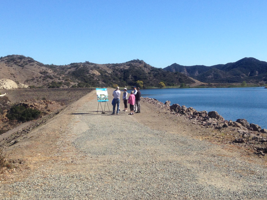 A small group of people standing by a lake, looking at displays on easels, with hills in the background.