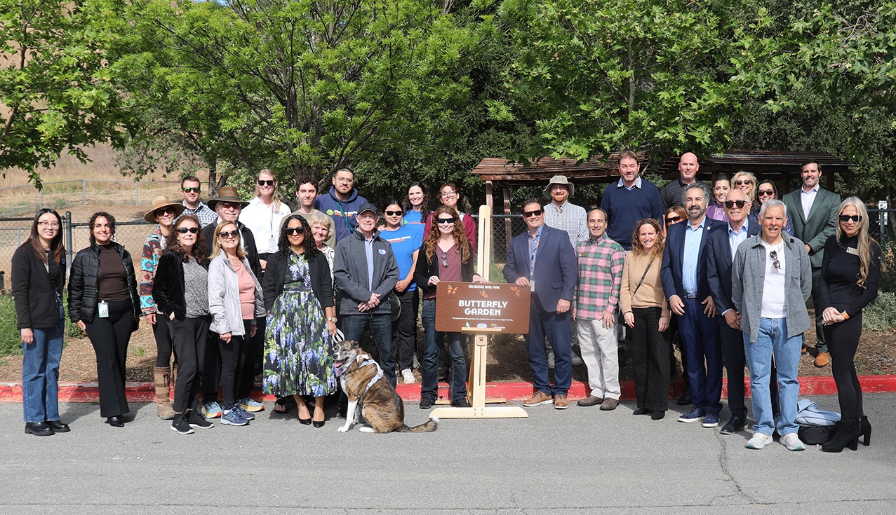 A diverse group of people gathered for a photo in front of a sign for a "Butterfly Garden." A dog is also present.