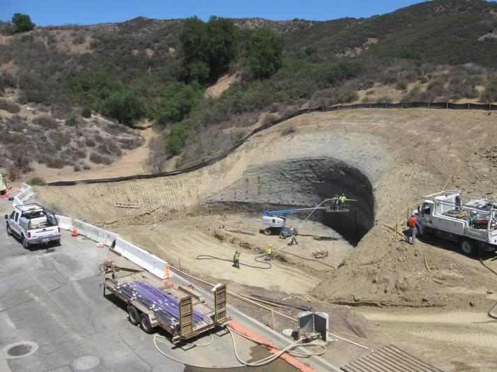 Construction site with vehicles, workers, and earth excavation surrounded by hills with sparse vegetation.