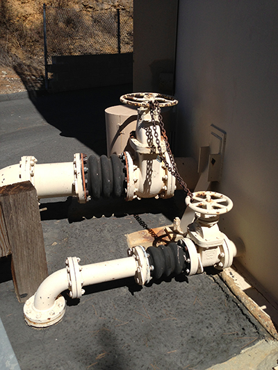 Pipes with valves and rubber joints, secured with a chain, are installed on the ground near a building.