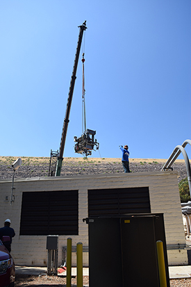 A crane lifting machinery onto a building rooftop with two people supervising.
