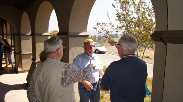 Three men talking under an archway, with trees and a road in the background.