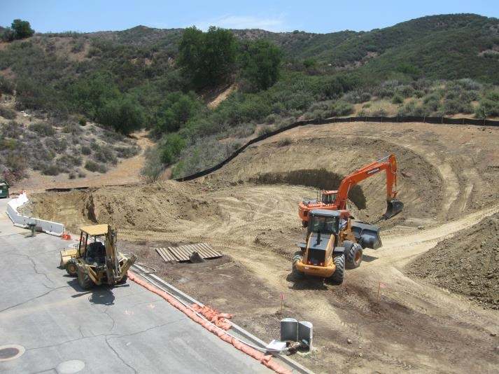 Construction site with excavator and bulldozer on a dirt area, surrounded by hills and vegetation.
