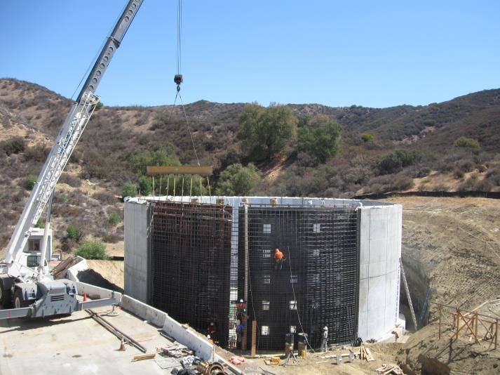 Construction site with a large crane lifting materials, workers inside a cylindrical structure made of steel rebar, in a hilly area.