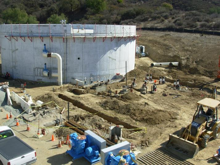 Construction site with a large circular concrete structure, machinery, and various building materials and pipes around.