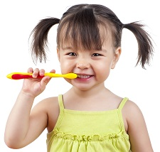 A young girl in a yellow dress is brushing her teeth with a red and yellow toothbrush, smiling.