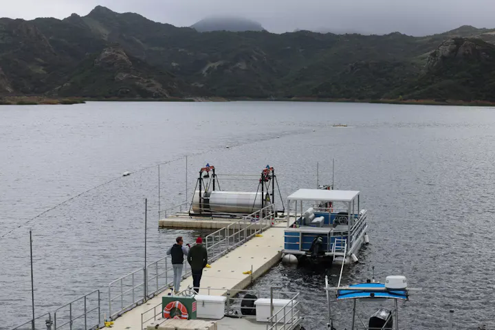 A serene lake scene with mountains in the background, a dock, a boat, and two people chatting near a cylindrical structure.