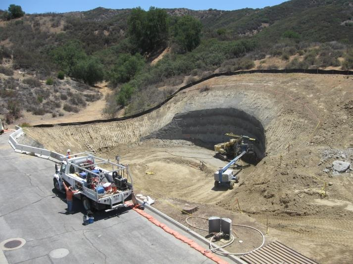 Construction site with excavation and equipment near a dirt slope, surrounded by dry vegetation and hills.