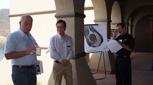 Three men standing near a poster with a diagram, two men holding papers, one holding a cup, outdoors near archways.