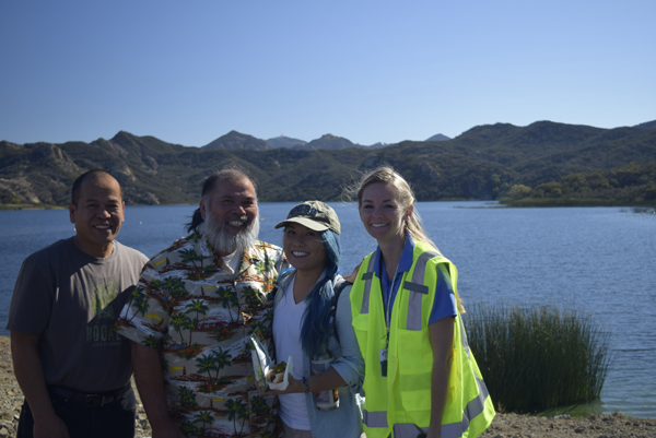 Four people posing by a lake with mountains in the background, one wearing a safety vest.