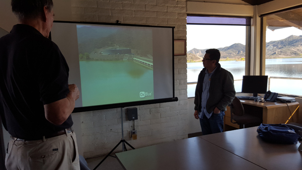 Two people in a room with a projector screen showing a dam or water structure, near a window with a lake view.