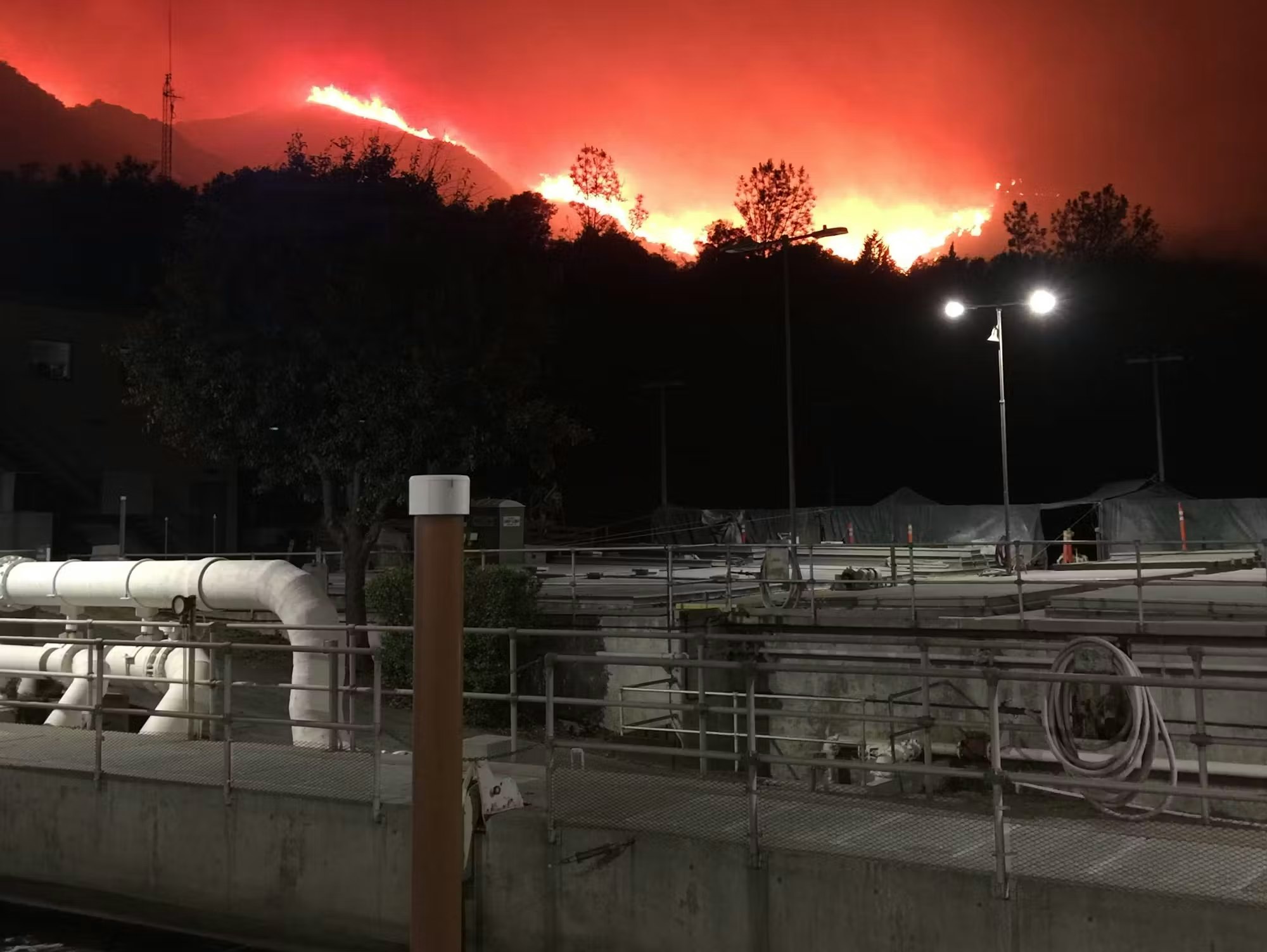 A dramatic nighttime scene of a wildfire raging on a hillside, with industrial structures and lights in the foreground.