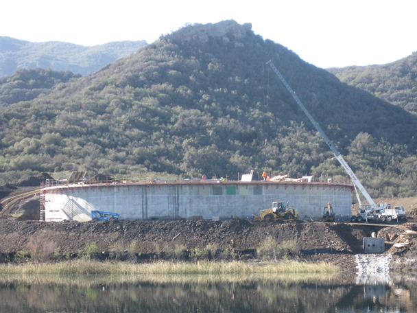 Construction site with a large concrete structure, cranes, and equipment, set against a backdrop of hills and reflective water.