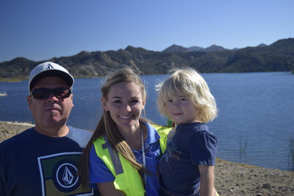 A man, woman, and child posing by a lake with mountains in the background; the woman is in a neon vest.