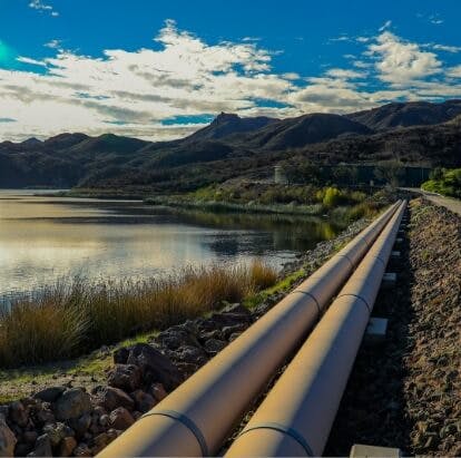 Pipelines running along a rocky lakeshore with mountains in the background and a partly cloudy sky.