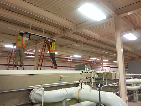 Two workers on ladders in a large industrial room with pipes and machinery, wearing safety vests and helmets.