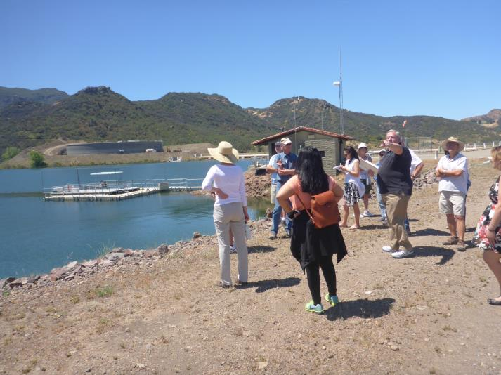 A group of people standing by a lake, with hills in the background, under a clear blue sky.