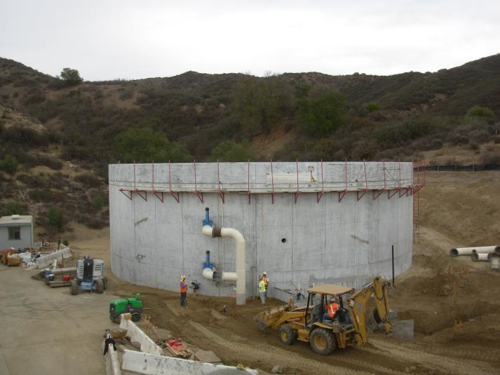 Construction site with a round concrete structure, pipes, workers, and construction machinery in a hilly area.