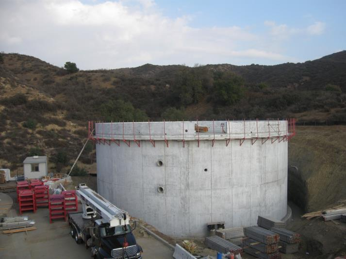 A large concrete cylindrical structure under construction, with scaffolding, surrounded by hills, equipment, and a truck nearby.