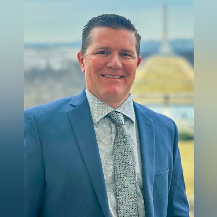 A smiling man in a blue suit and patterned tie stands outdoors with a view of a dome in the background.