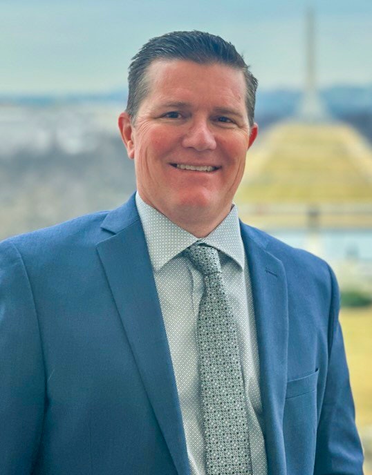 A smiling man in a blue suit and patterned tie stands outdoors with a view of a dome in the background.