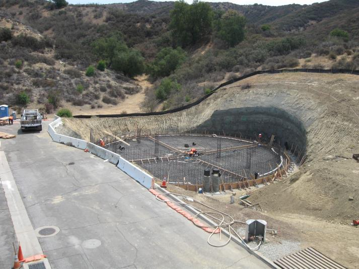 Construction site on a hillside with steel rebar framing for a circular foundation, surrounded by vegetation and equipment.