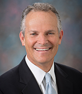 A person in a suit smiling, set against a blurred background.